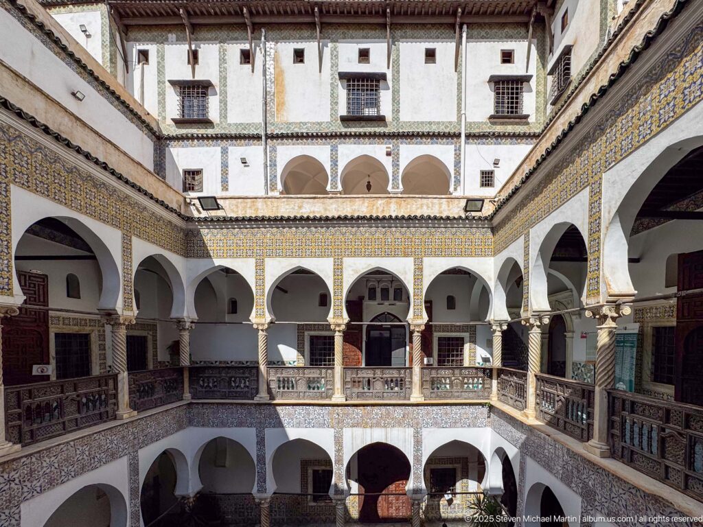 Elevated courtyard view with stacked white arcades, wooden railings, patterned tile bands, upper walls, and strong sunlight and shadow.