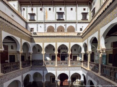 Elevated courtyard view with stacked white arcades, wooden railings, patterned tile bands, upper walls, and strong sunlight and shadow.