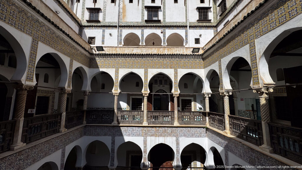 Elevated frontal view across the courtyard toward stacked white arcades, dark wooden railings, and blue-and-yellow tile bands.
