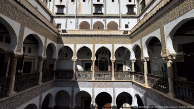 Elevated frontal view across the courtyard toward stacked white arcades, dark wooden railings, and blue-and-yellow tile bands.