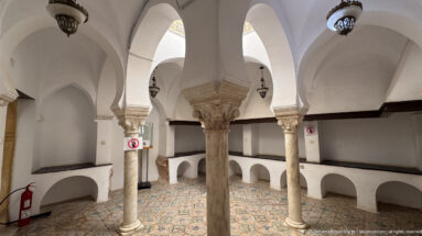 White vaulted room with pale stone columns, carved capitals, built-in wall benches, patterned tile floor, and hanging lamps.