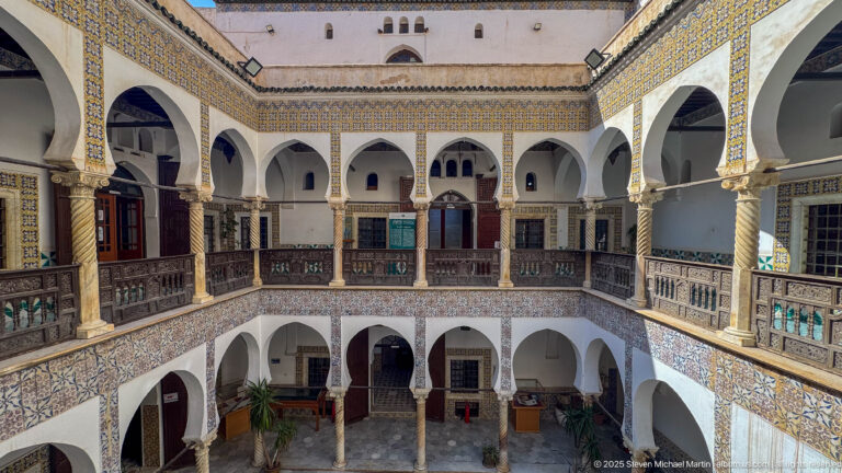 Oblique elevated courtyard view with white arches, spiral columns, patterned tilework, dark wooden galleries, and lower court visible below.
