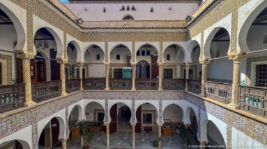 Oblique elevated courtyard view with white arches, spiral columns, patterned tilework, dark wooden galleries, and lower court visible below.