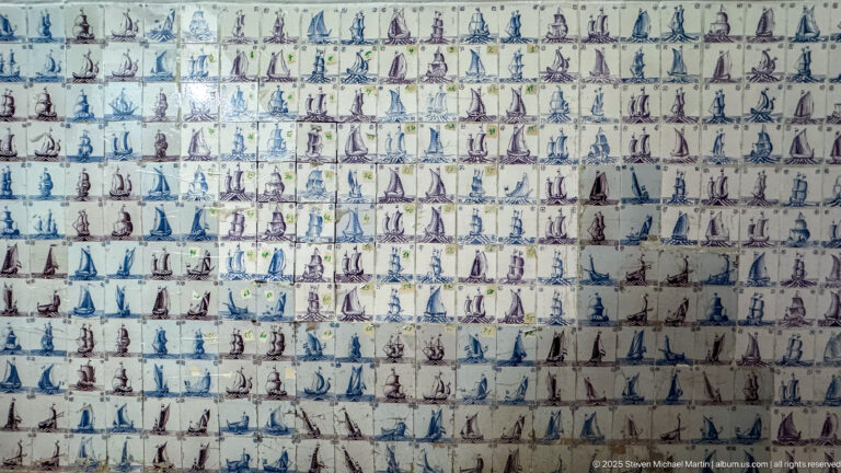 Large wall covered with rows of small ceramic tiles depicting varied sailing ships in blue, purple, and brown.