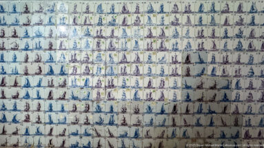 Large wall covered with rows of small ceramic tiles depicting varied sailing ships in blue, purple, and brown.