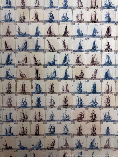 Close wall view of blue and purple sailing-ship tiles with varied sail plans, cracked glaze, stained grout, and surface wear.