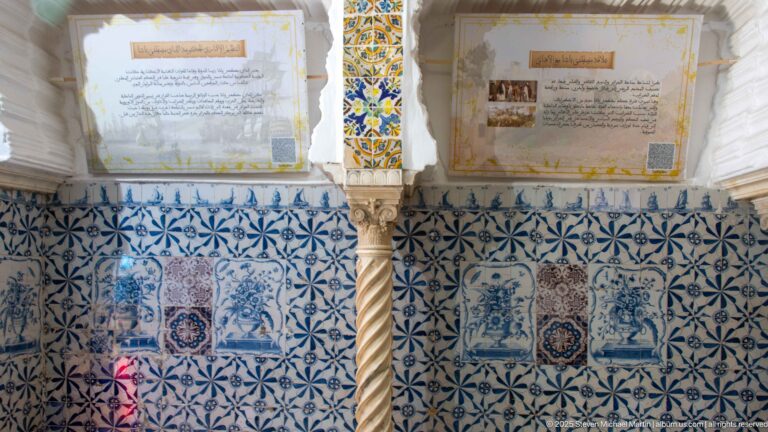 Interior wall covered in blue-and-white tiles, with a spiral stone column and Arabic interpretive panels mounted above.