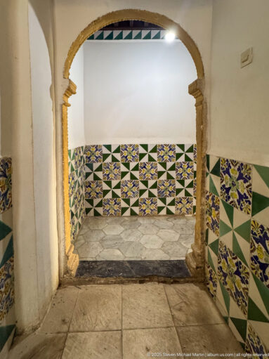 Scalloped pale stone doorway opening into a small white room lined with green, blue, yellow, and white geometric tiles.