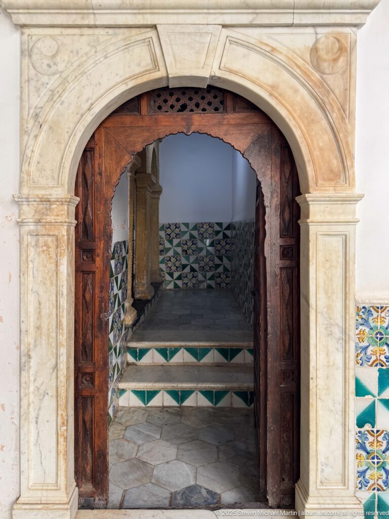 Stone arched portal with open dark wooden doors leading to short steps and green-and-white geometric tiled walls.