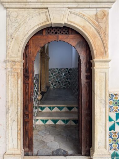 Stone arched portal with open dark wooden doors leading to short steps and green-and-white geometric tiled walls.