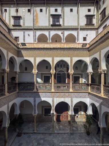 Wide elevated view of the palace courtyard with multiple arcade levels, dark wooden galleries, patterned tile bands, and upper windows.
