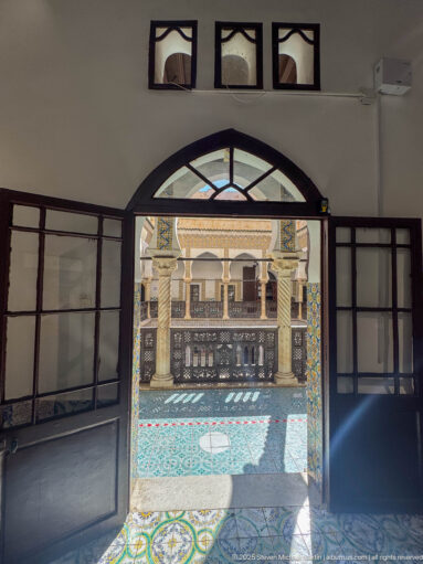 Open dark glazed doors framing a sunlit palace gallery with carved railing, spiral columns, patterned floor, and courtyard beyond.