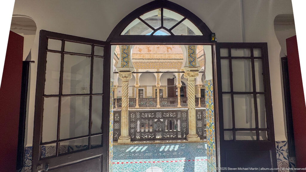 Open glazed doors beneath an arched transom, framing spiral columns, carved wooden railing, and tiled courtyard architecture.