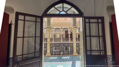Open glazed doors beneath an arched transom, framing spiral columns, carved wooden railing, and tiled courtyard architecture.