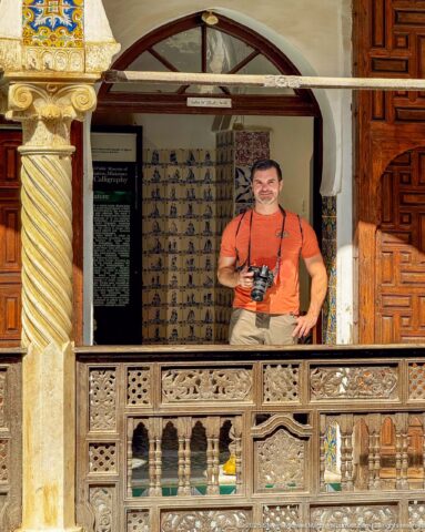 Man in an orange shirt holding a camera behind a carved wooden railing, beside a spiral column and geometric wooden door.