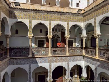 Wide courtyard view with a visitor in orange centered behind the upper wooden gallery railing among white arches and patterned tiles.
