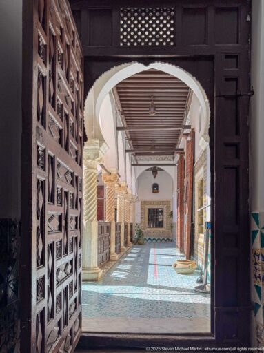 Dark carved doorway framing a long sunlit gallery with spiral columns, wooden railings, patterned floor tiles, and a red guide line.