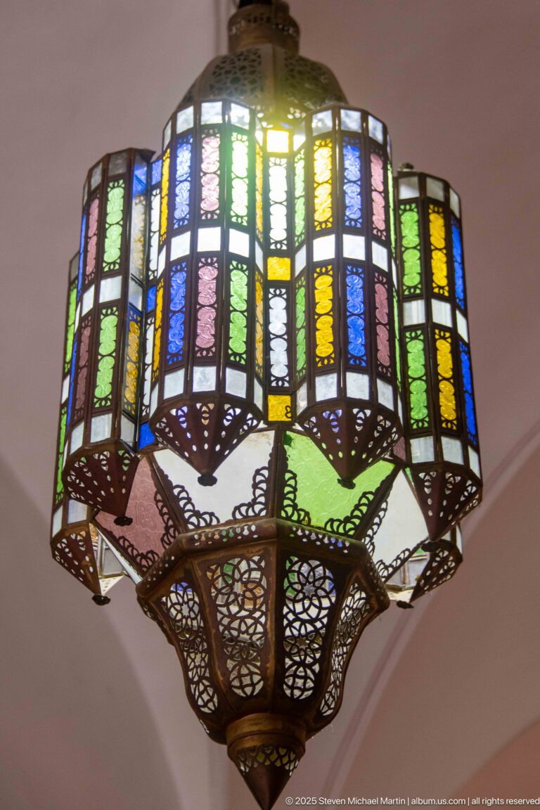 Large suspended lantern with multicolored textured glass panes set in an ornate dark metal framework and pierced lower basket.