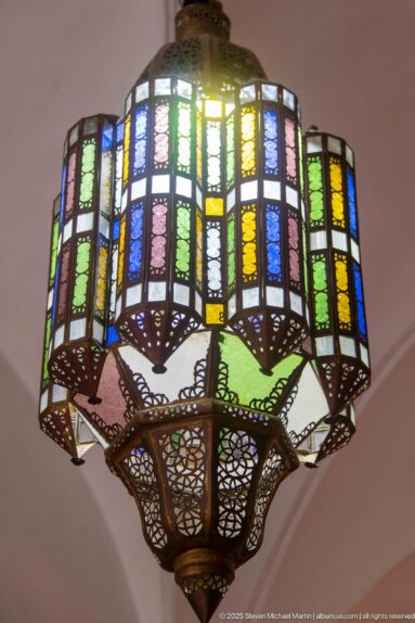 Large suspended lantern with multicolored textured glass panes set in an ornate dark metal framework and pierced lower basket.