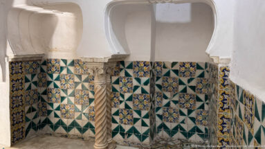 Spiral-fluted stone column before green-and-white geometric wall tiles, blue-and-yellow floral squares, and a white plaster arch.