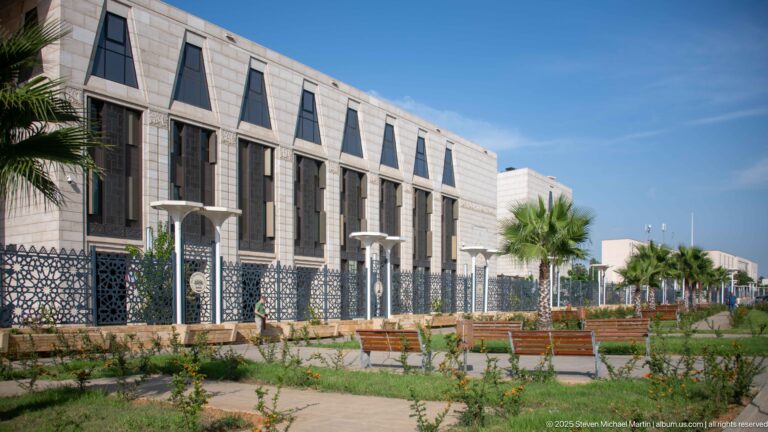 Landscaped walkway with palms and benches beside the pale-stone exterior of the Great Mosque of Algiers.
