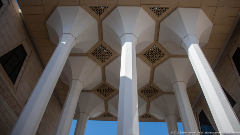 Upward view of faceted white column heads meeting a ceiling punctuated by square geometric grilles.