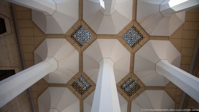 Symmetrical upward view of four flared white supports around square patterned grilles in the mosque portico ceiling.