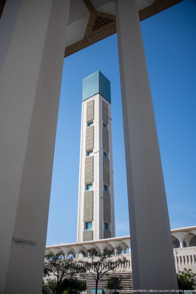 Square minaret with blue-green crown framed between large white portico columns under a clear sky.