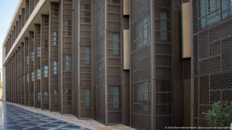 Long mosque facade lined with tall perforated metal screens over recessed glazing beside a paved walkway.