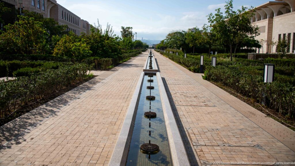 Long narrow water channel centered in a brick walkway with hedges and trees in the mosque grounds.