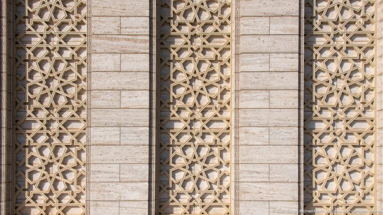 Three tall geometric lattice panels set into a pale horizontally jointed stone mosque wall.