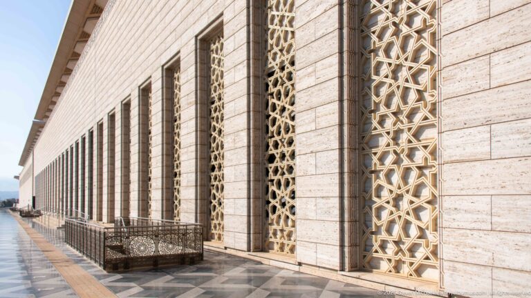 Long pale-stone mosque facade with repeated tall geometric lattice panels receding beside dark paving.