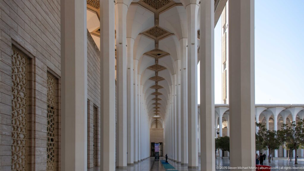 Long covered mosque colonnade with repeated white columns, screened wall openings, and alternating sunlight and shade.
