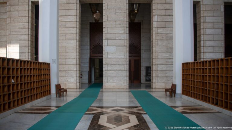 Wooden shoe racks flank green carpet runners leading toward tall interior doorways in the mosque.