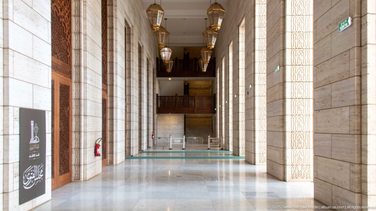 Tall mosque corridor with pale stone piers, hanging lanterns, wooden doors, green runners, and polished floor.