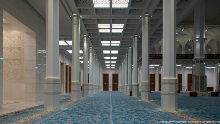 Large mosque prayer hall with rows of white columns, blue patterned carpet, ceiling bays, and pendant lights.