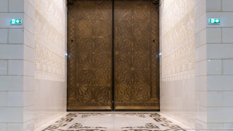 Tall dark carved double doors framed by pale patterned walls and geometric stone flooring inside the mosque.