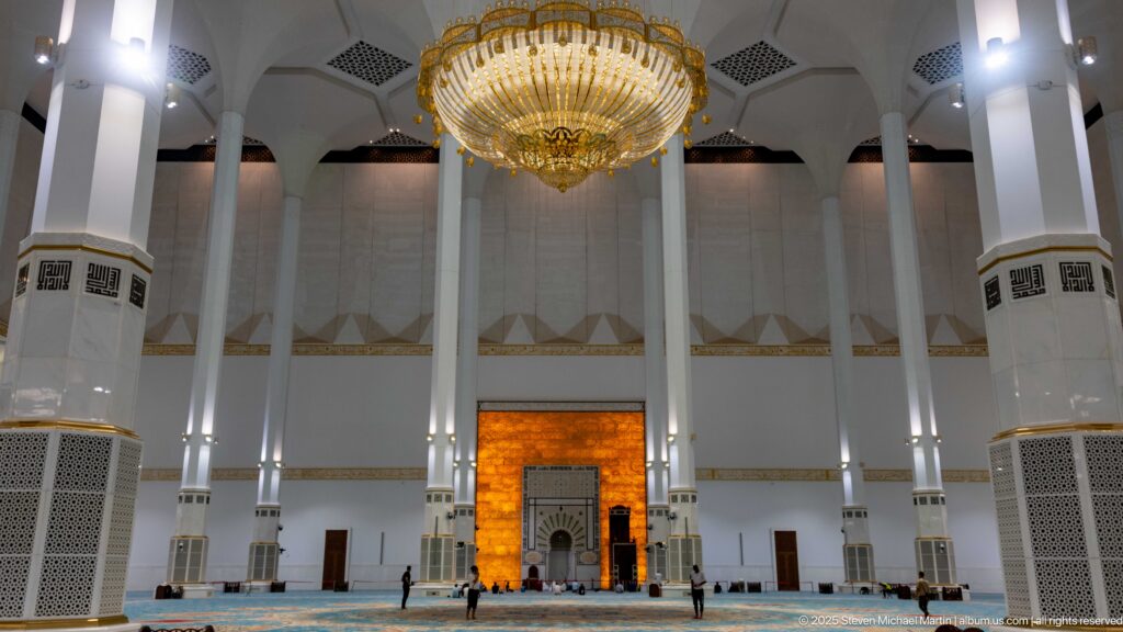Wide prayer hall with blue carpet, white columns, large chandelier, and illuminated mihrab wall at the far end.