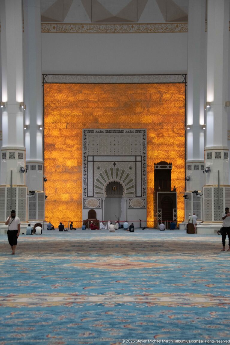 Illuminated amber qibla wall with central mihrab, dark pulpit-like furnishing, and worshippers on blue carpet.