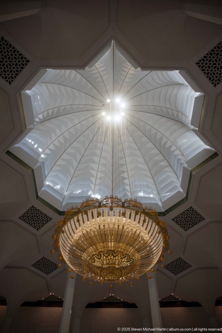 Upward view of a chandelier beneath a faceted star-shaped ceiling recess with geometric ventilation grilles.