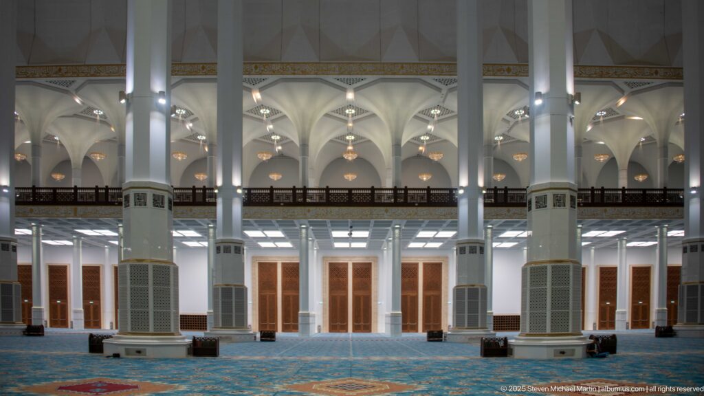 Wide mosque prayer hall with white columns, blue carpet, hanging lights, and a dark gallery across the rear.