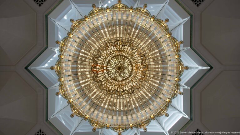 Close upward view of a circular gilded chandelier with radiating glass elements inside a star-shaped white ceiling recess.