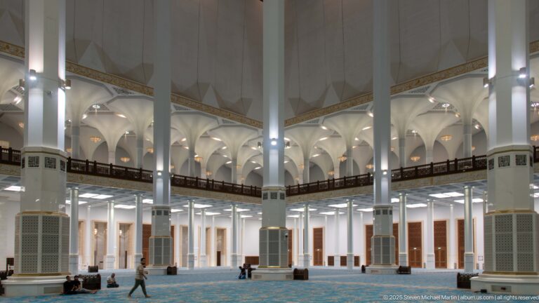 White columns and an upper wooden gallery rise above a turquoise patterned carpet with several people scattered across the prayer hall.