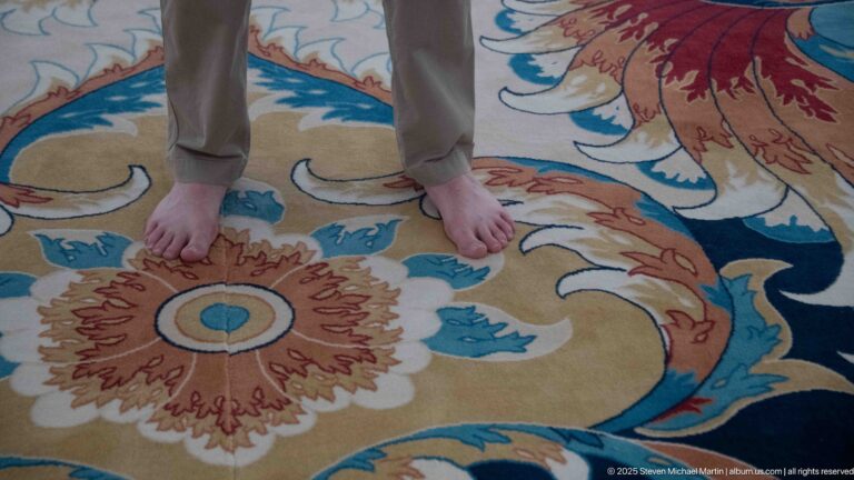 Bare feet stand on a blue, cream, and rust floral carpet inside the mosque.