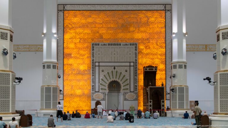 Worshippers sit and stand before the mihrab and glowing amber qibla wall between tall white columns.