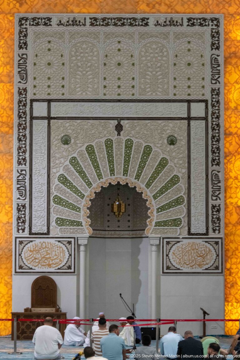 Decorated mihrab with green and white radiating ornament above worshippers seated on the carpet.