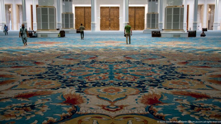 Large blue floral carpet extends toward white columns and wooden doors, with a few people walking across the hall.