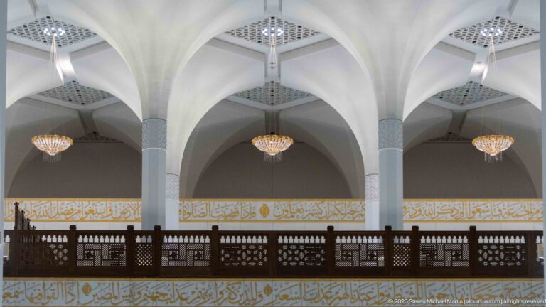 Dark wooden gallery spans between pale columns and branching arches, with chandeliers and gold calligraphic bands behind.