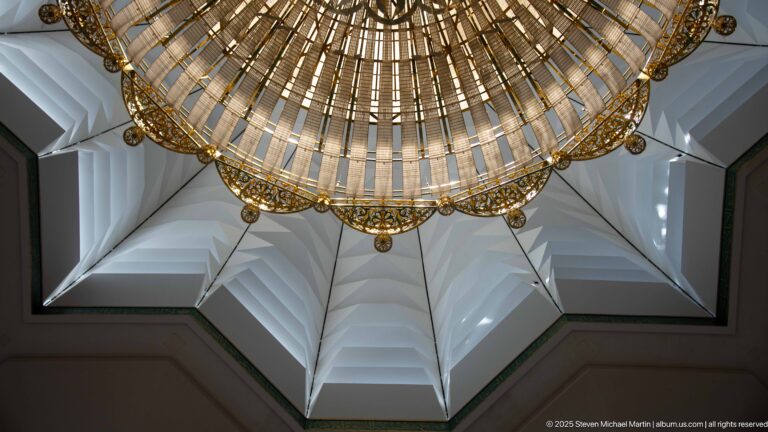Large circular chandelier hangs beneath a faceted white ceiling recess viewed directly from below.