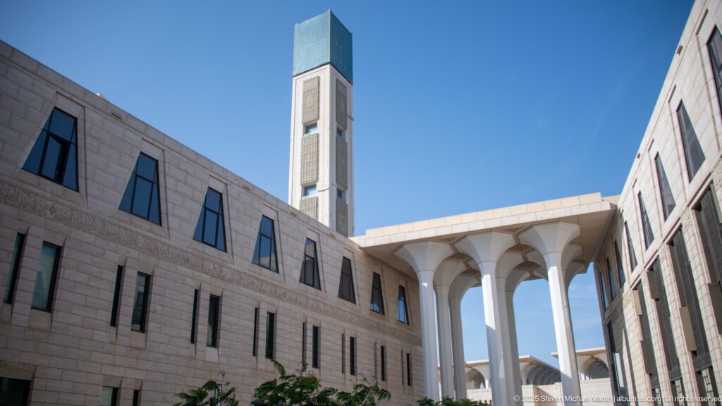 Square minaret with blue-green crown rising above stone buildings and a white colonnade at the Great Mosque of Algiers.